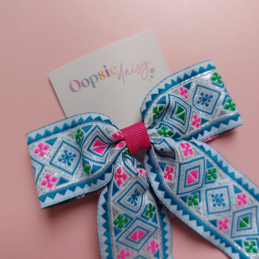 A hair bow with a geometric pattern in blue, pink, green, and white displayed on a pink background with a card that reads 'Oopsie Daisy'.