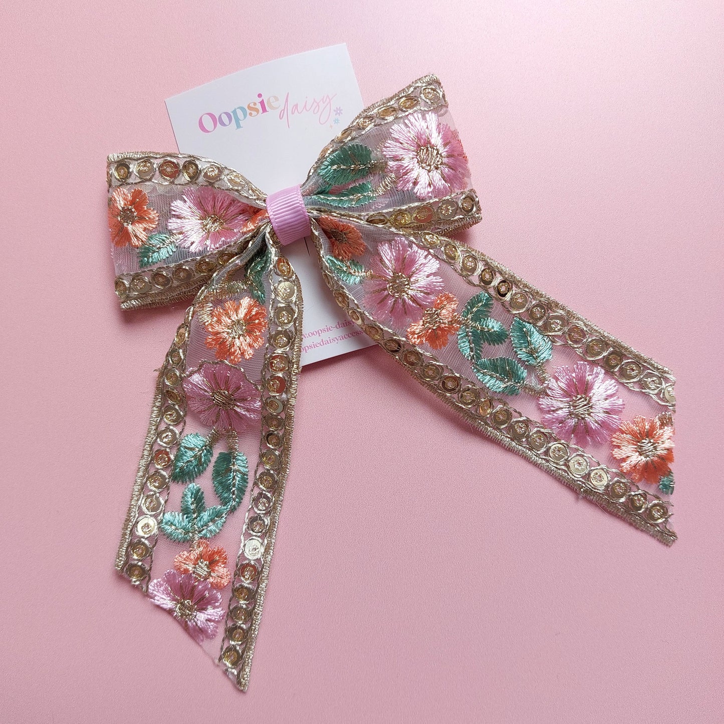A floral embroidered hair bow with pink, orange, and green flowers, gold sequined trim, and a pink ribbon center, displayed on a pink background with a card that reads 'Oopsie Daisy'.