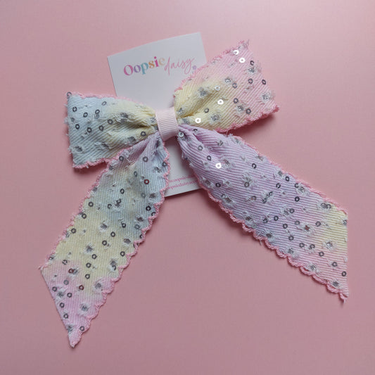 A pastel hair bow with sequins, featuring a light purple, yellow, and pink gradient pattern with silver sequins and scalloped edges, displayed on a pink background with a brand tag.