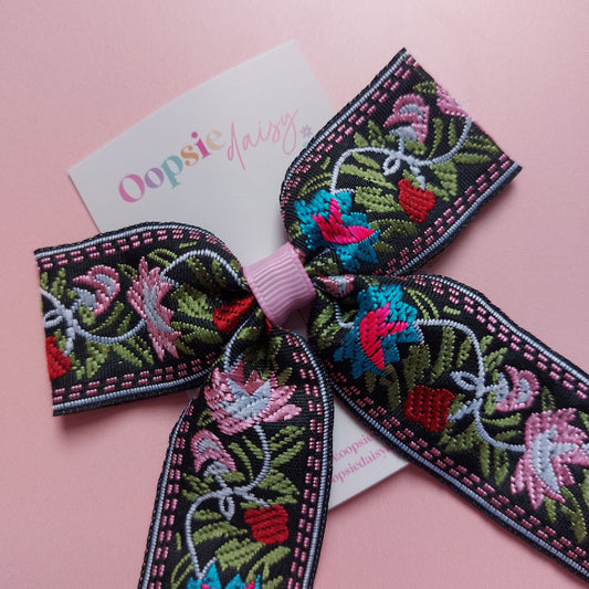A decorative embroidered bow with a pink center band, laid on a soft pink background. The bow features floral embroidery in pink, turquoise, red, green, and white on a black base.