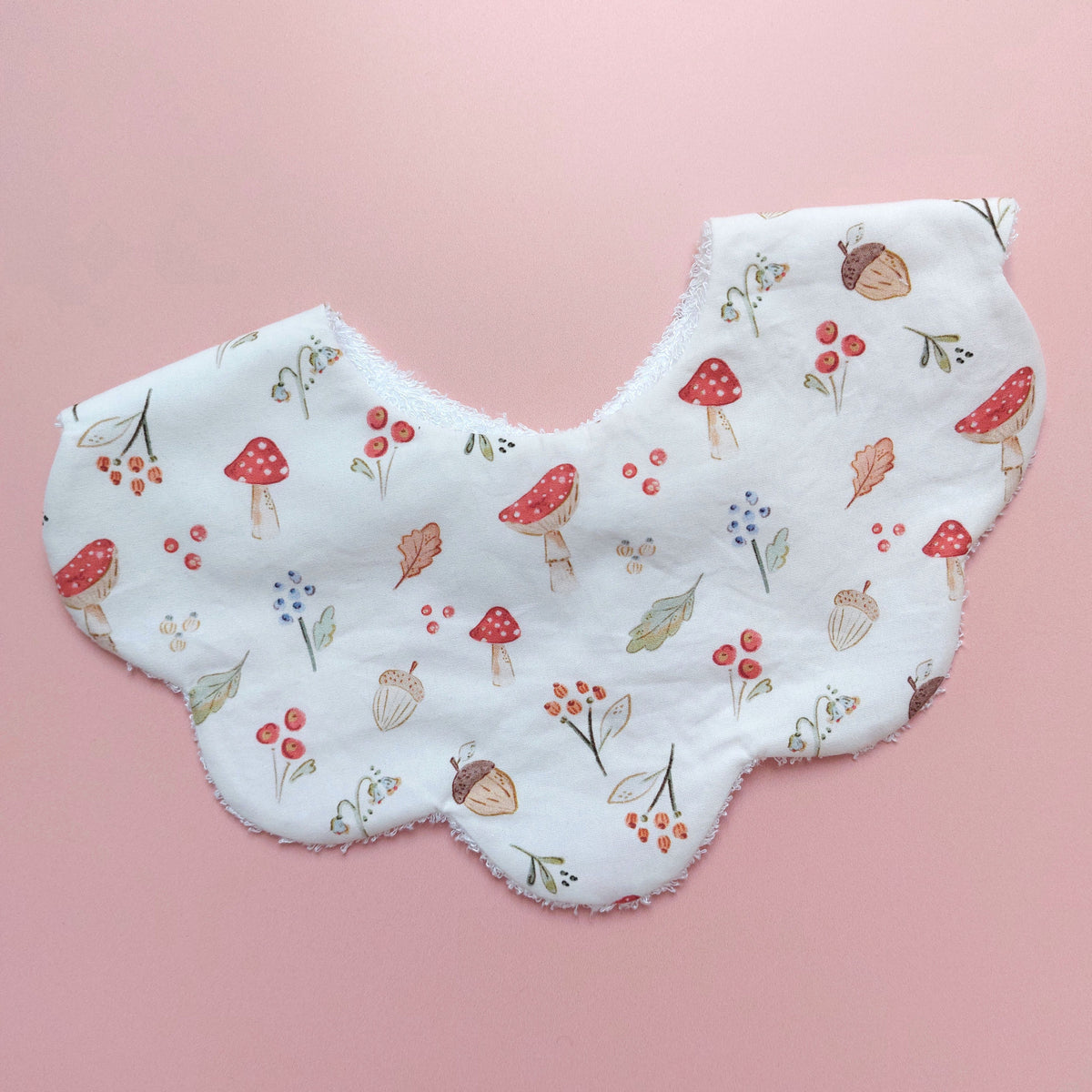 Scallop edged baby bib in white with autumnal pattern. Featuring mushrooms, acorns, leaves and autumnal flowers.