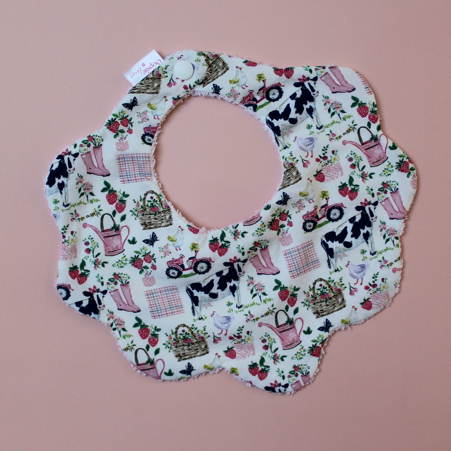 Scallop baby bib with girls farm themed pattern. Cream background with cows, pink watering cans, tractors and strawberries.