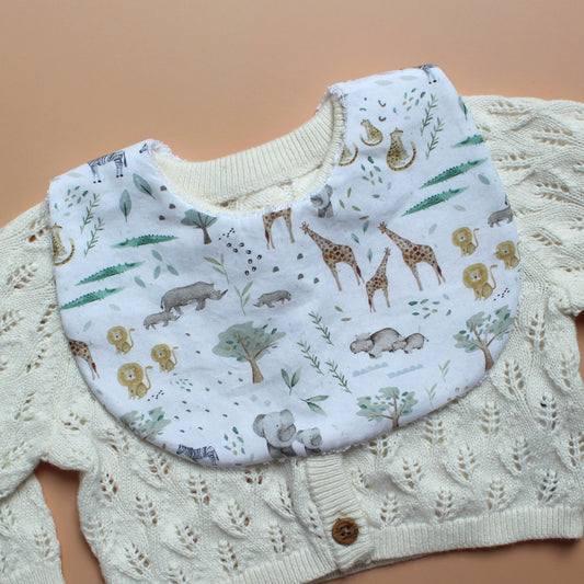 A baby bib with an animal safari print, styled on top of a cream-colored cable-knit cardigan against a solid peach background.