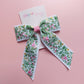 A hair bow with a white background and an embroidered floral pattern in pink, green, and yellow. 