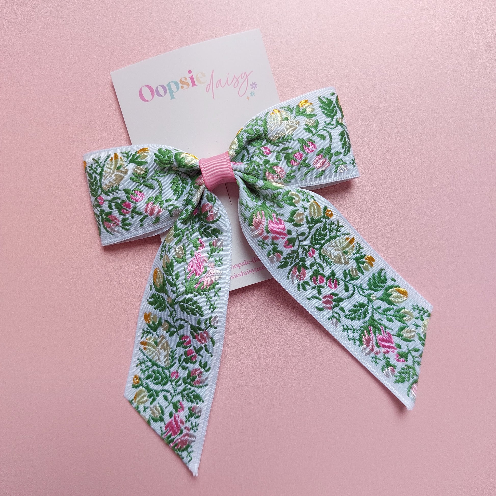 A hair bow with a white background and an embroidered floral pattern in pink, green, and yellow. 