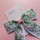 A hair bow with a white background and an embroidered floral pattern in pink, green, and yellow. 