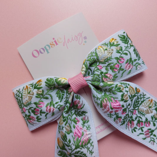 A hair bow with a white background and an embroidered floral pattern in pink, green, and yellow. 