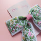 A hair bow with a white background and an embroidered floral pattern in pink, green, and yellow. 