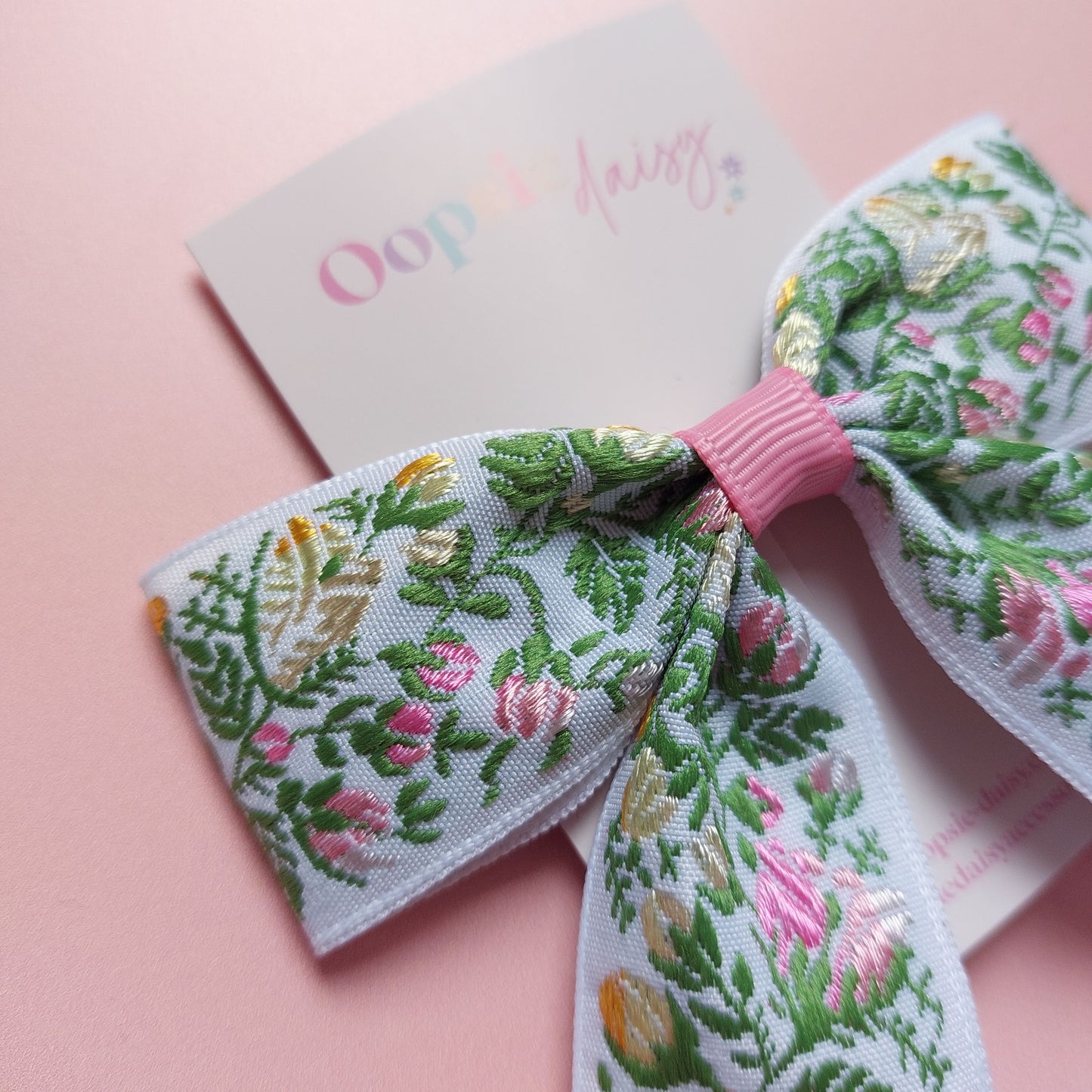 A hair bow with a white background and an embroidered floral pattern in pink, green, and yellow. 
