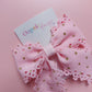 A pink ribbon hair bow with scalloped edges and small gold glitter dots, attached to a white card printed with the brand name 'Oopsie Daisy'.