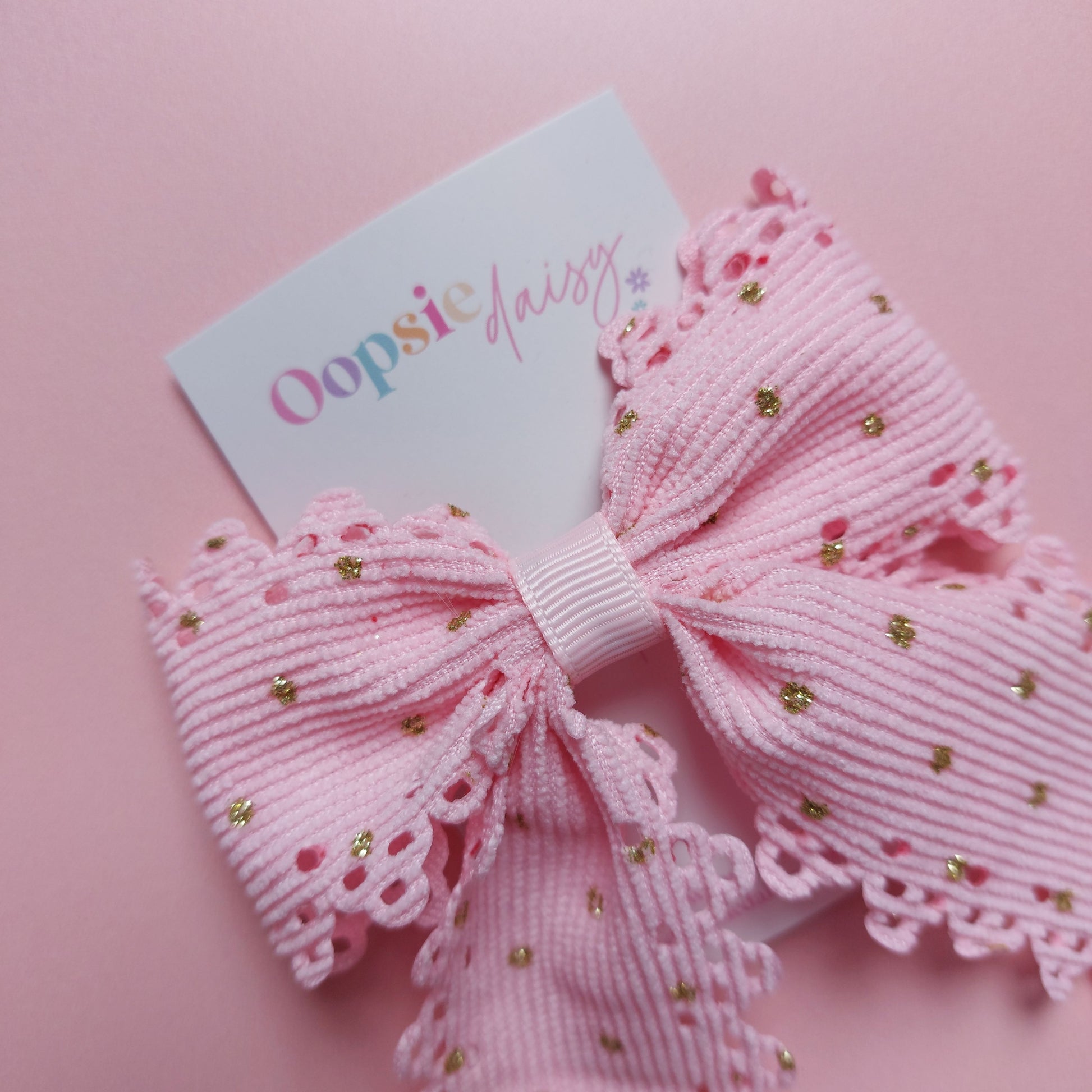A pink ribbon hair bow with scalloped edges and small gold glitter dots, attached to a white card printed with the brand name 'Oopsie Daisy'.