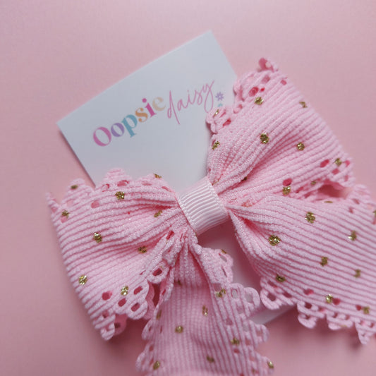 A pink ribbon hair bow with scalloped edges and small gold glitter dots, attached to a white card printed with the brand name 'Oopsie Daisy'.