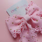 A pink ribbon hair bow with scalloped edges and small gold glitter dots, attached to a white card printed with the brand name 'Oopsie Daisy'.