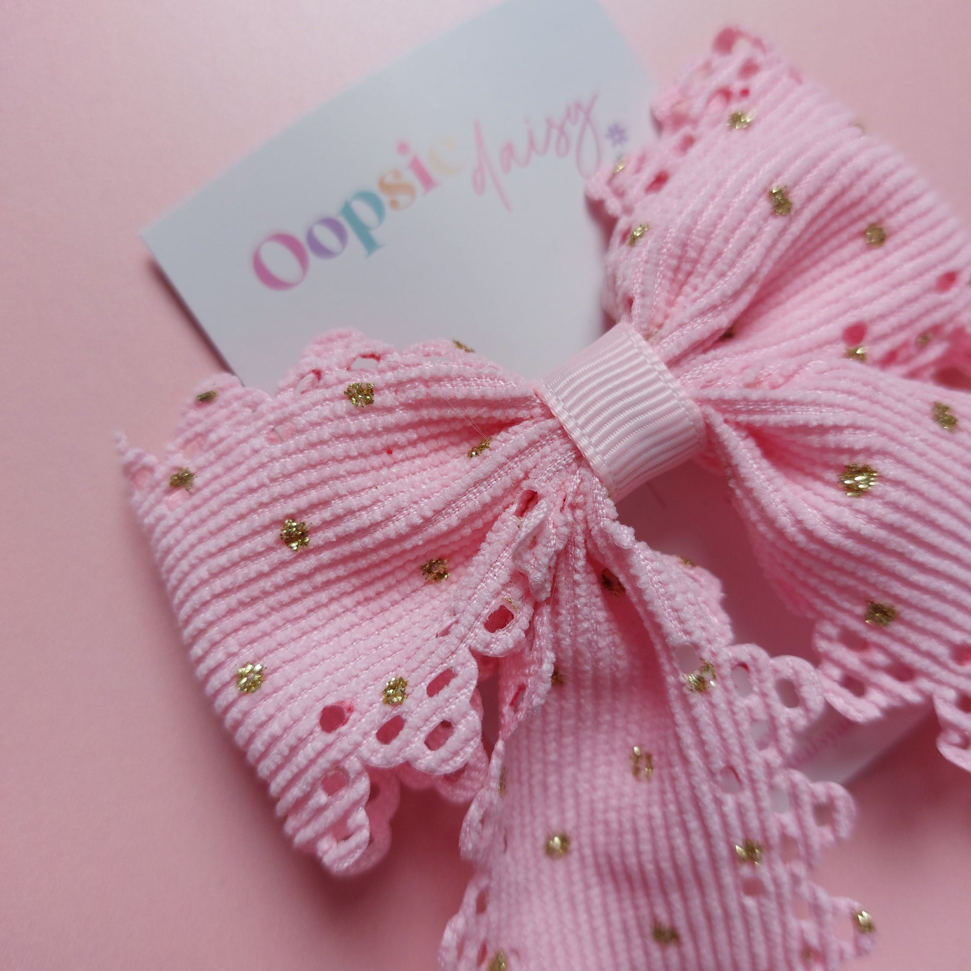 A pink ribbon hair bow with scalloped edges and small gold glitter dots, attached to a white card printed with the brand name 'Oopsie Daisy'.