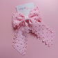 A pink ribbon hair bow with scalloped edges and small gold glitter dots, attached to a white card printed with the brand name 'Oopsie Daisy'.
