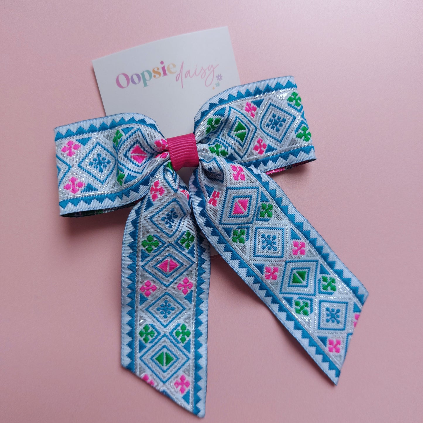 A hair bow with a colourful geometric pattern in blue, pink, green, and white, attached to a white card that reads 'Oopsie Daisy' and displayed on a pink background.