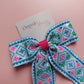 A hair bow with a geometric pattern in blue, pink, green, and white displayed on a pink background with a card that reads 'Oopsie Daisy'.