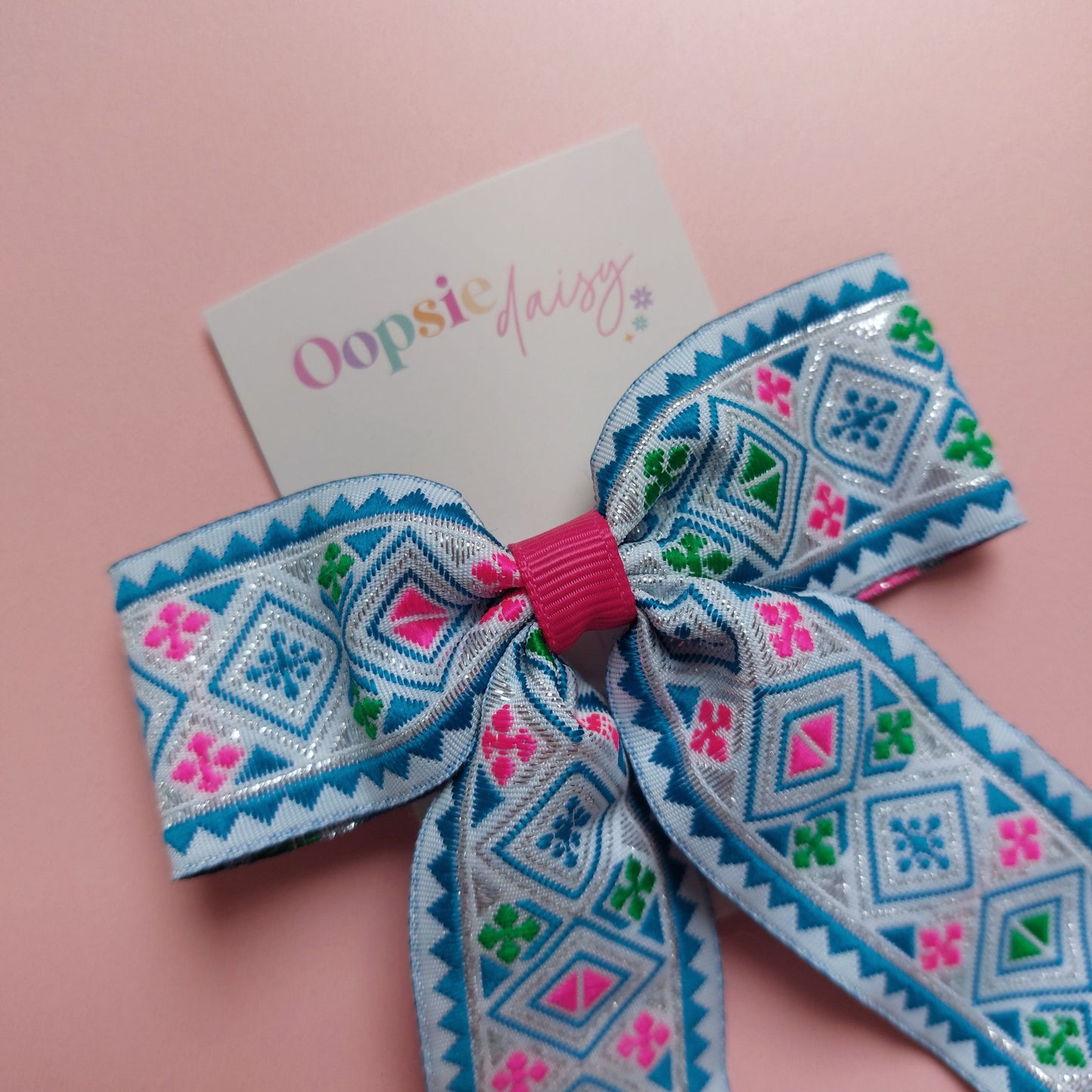 A hair bow with a geometric pattern in blue, pink, green, and white displayed on a pink background with a card that reads 'Oopsie Daisy'.