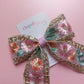A floral embroidered hair bow with pink, orange, and green flowers, gold sequined trim, and a pink ribbon center, displayed on a pink background with a card that reads 'Oopsie Daisy'.
