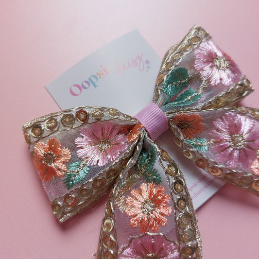 An embroidered hair bow with pink, orange, and green floral details, gold sequins, and sheer mesh ribbon, shown on a pink background with a branded card.