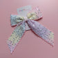 A pastel hair bow with sequins, featuring a light purple, yellow, and pink gradient pattern with silver sequins and scalloped edges, displayed on a pink background with a brand tag.