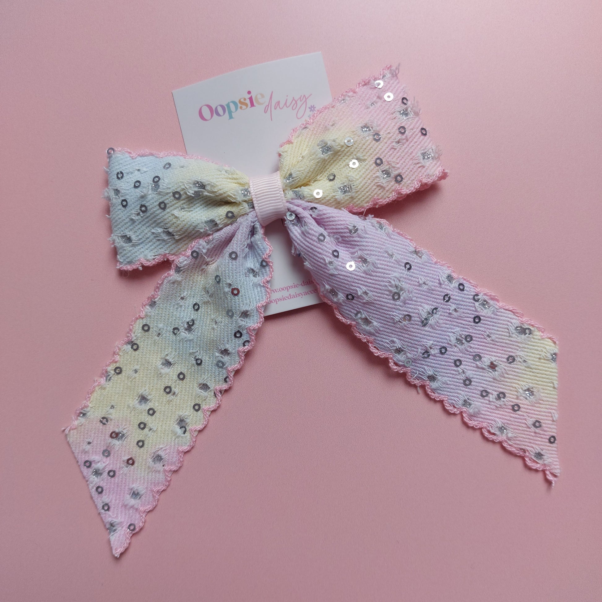 A pastel hair bow with sequins, featuring a light purple, yellow, and pink gradient pattern with silver sequins and scalloped edges, displayed on a pink background with a brand tag.