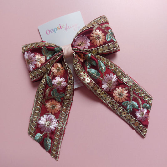 A decorative hair bow with intricate floral embroidery in pink, green, and gold sequins on a burgundy base, displayed on a soft pink background with a small brand tag.