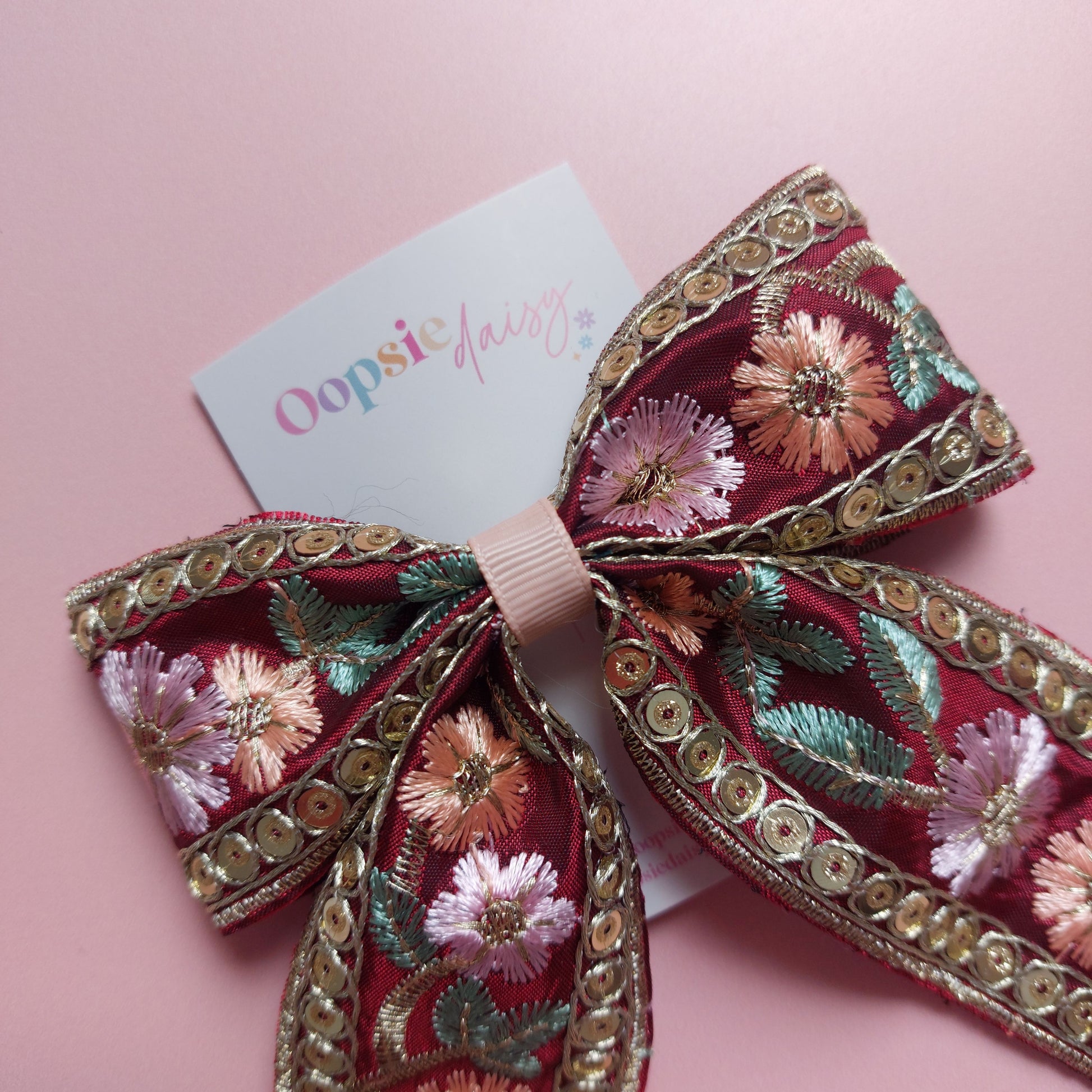 A large decorative hair bow made of burgundy fabric with embroidered pink and peach flowers, green leaves, and gold sequined trim, attached to a white card that reads 'Oopsie Daisy'.