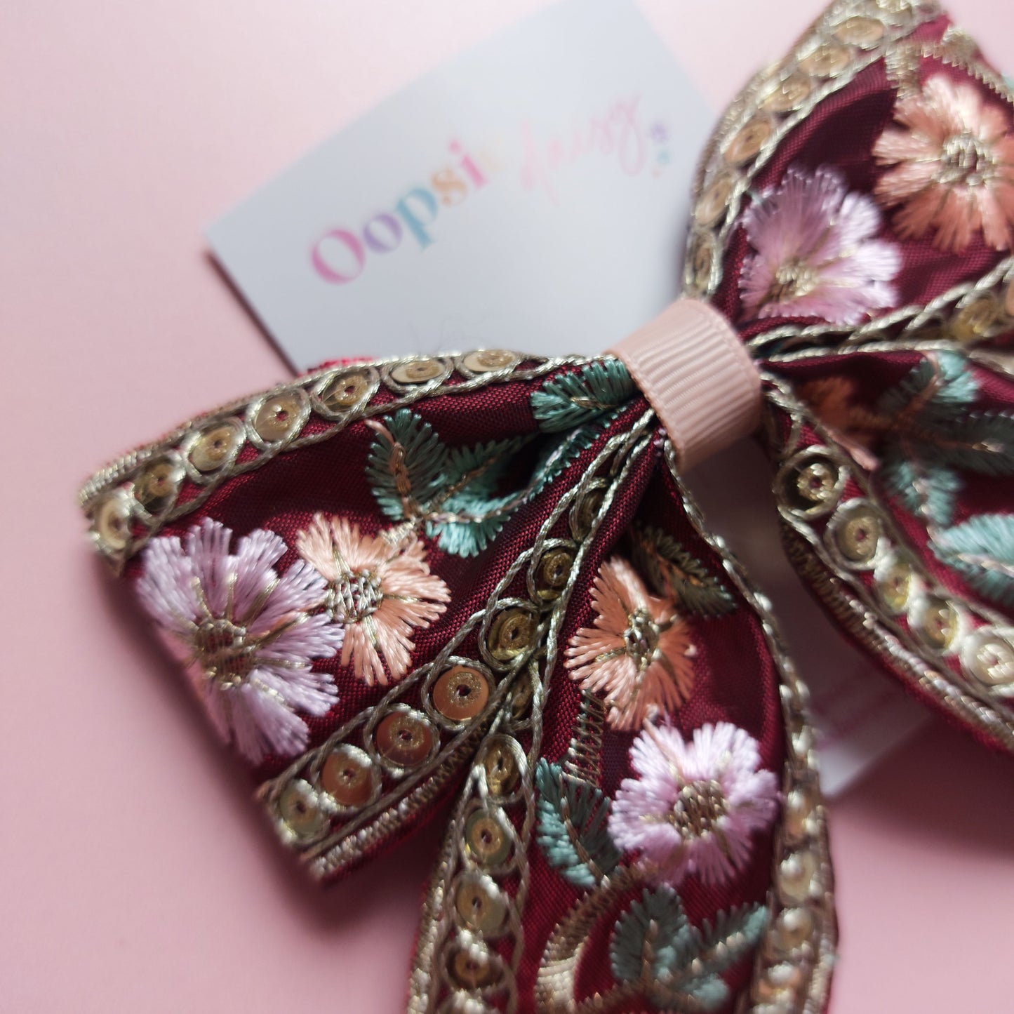 A decorative hair bow with intricate floral embroidery in pink, green, and gold sequins on a burgundy base, displayed on a soft pink background with a small brand tag.