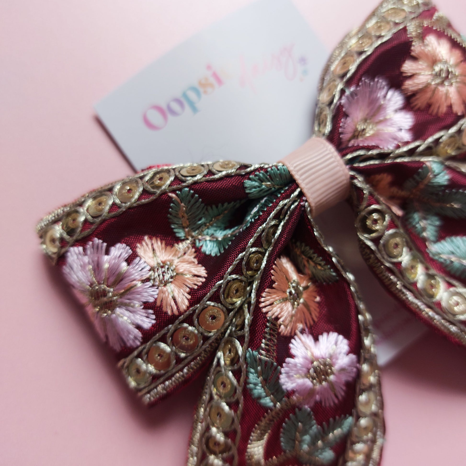 A decorative hair bow with intricate floral embroidery in pink, green, and gold sequins on a burgundy base, displayed on a soft pink background with a small brand tag.