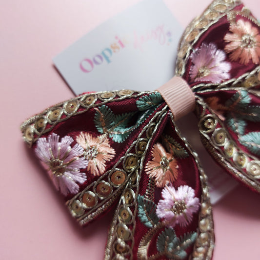 A decorative hair bow with intricate floral embroidery in pink, green, and gold sequins on a burgundy base, displayed on a soft pink background with a small brand tag.