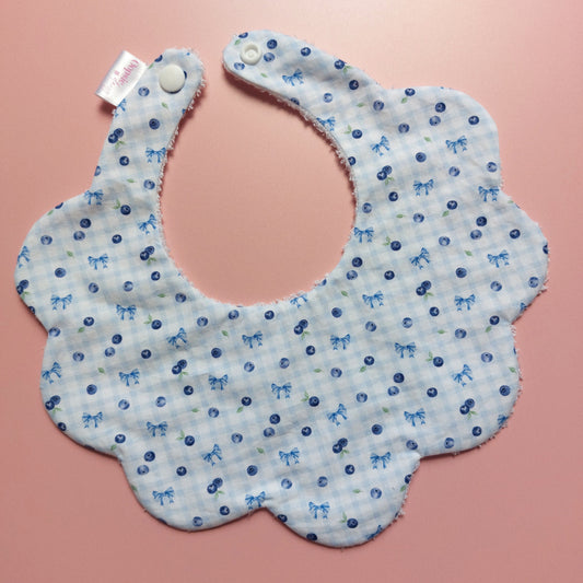 A scalloped fabric bib with a light blue and white gingham pattern, decorated with small blue bows and blueberries, displayed on a pink background.