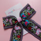 A decorative embroidered bow with a pink center band, laid on a soft pink background. The bow features floral embroidery in pink, turquoise, red, green, and white on a black base.