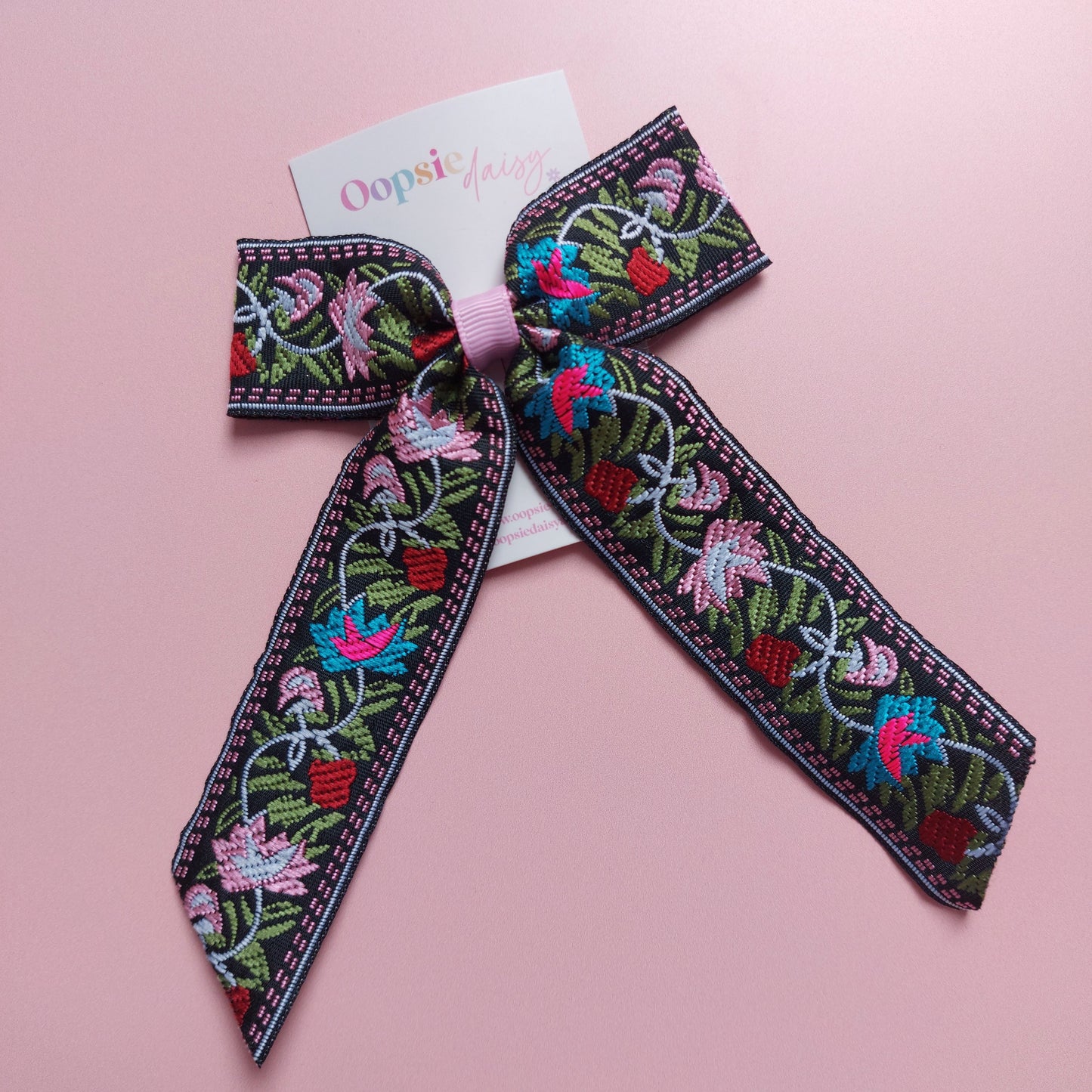 A decorative embroidered hair bow with a black base and colourful floral embroidery in pink, blue, green, and red.