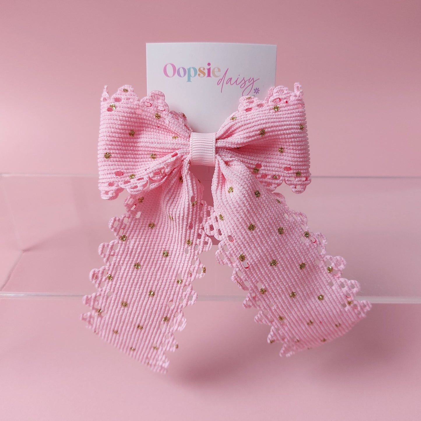 A pink ribbon bow with scalloped edges and small gold polka dots, attached to a white card printed with the brand name 'Oopsie Daisy', set against a solid pink background.