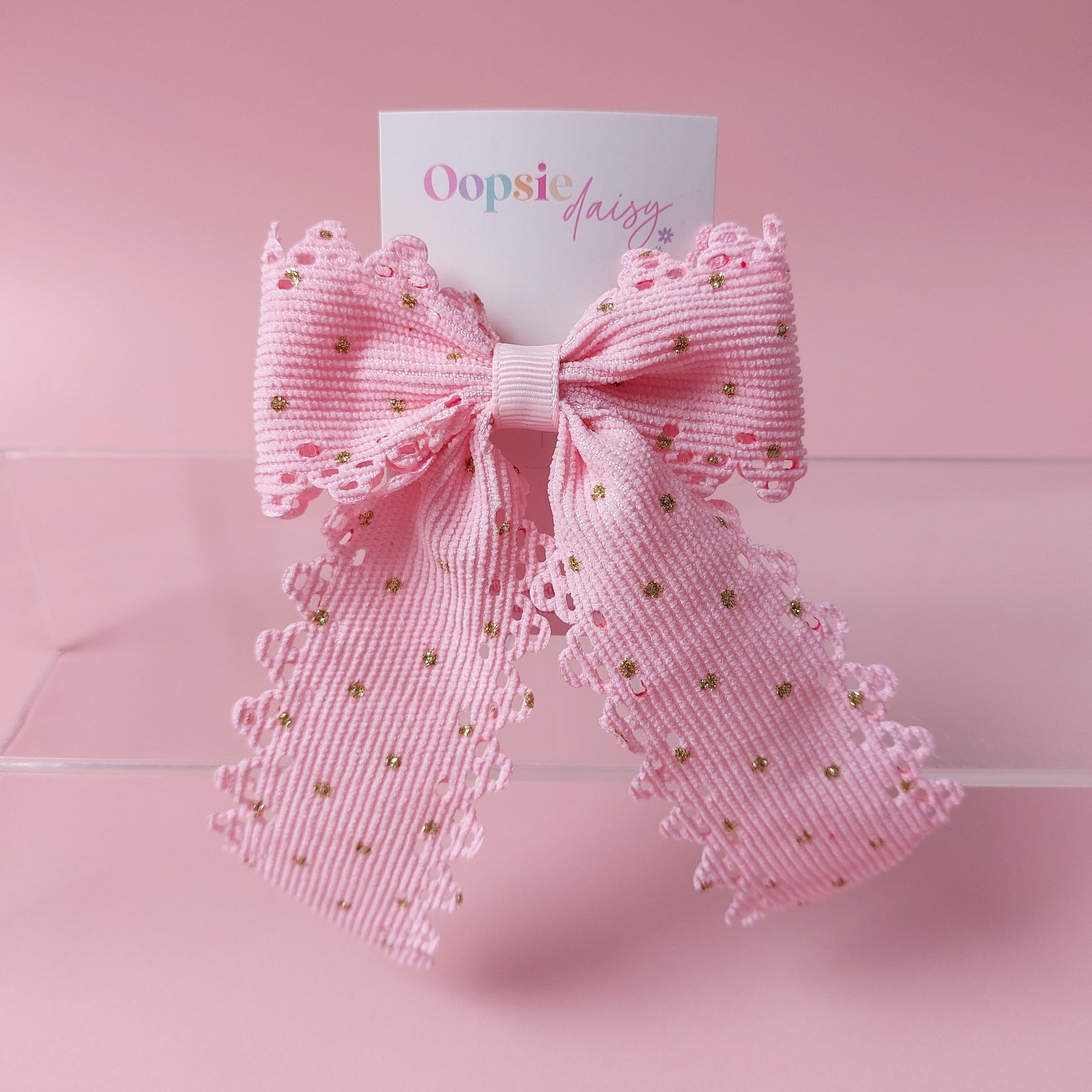 A pink ribbon bow with scalloped edges and small gold polka dots, attached to a white card printed with the brand name 'Oopsie Daisy', set against a solid pink background.