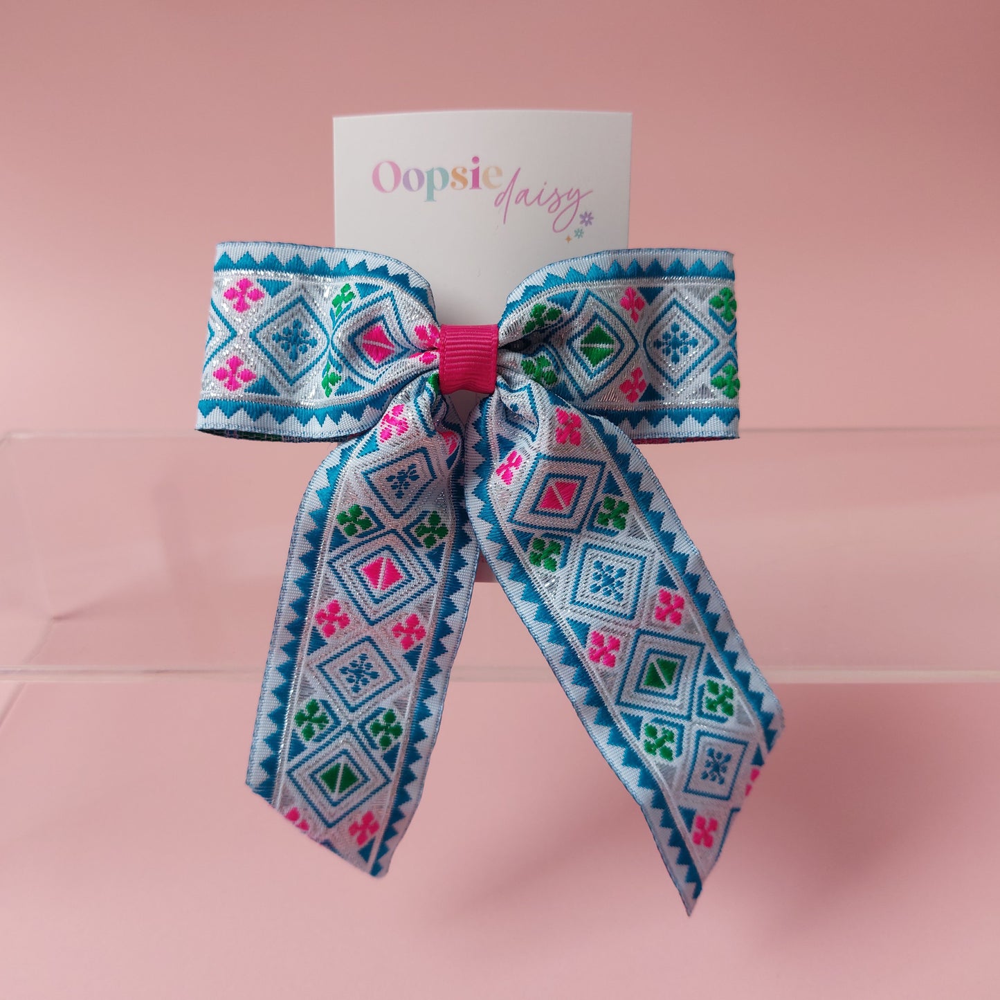 A hair bow with a colourful geometric pattern in blue, pink, green, and white, attached to a white card that reads 'Oopsie Daisy' and displayed on a pink background.