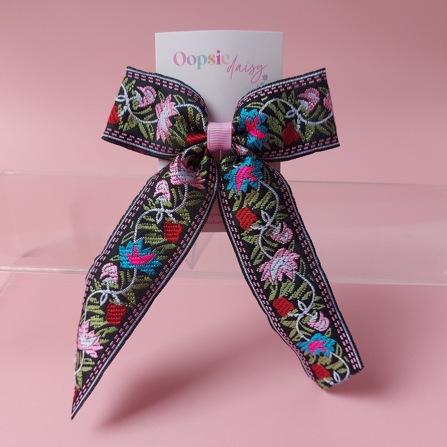 A decorative hair bow with a black embroidered floral pattern, pink ribbon center, and a backing card reading 'Oopsie dairy', photographed on a soft pink background.