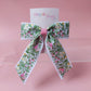 A floral hair bow with green leaves and pink flowers on a white ribbon base, tied with a pink center band, displayed on a pink card that reads 'Oopsie Daisy'.