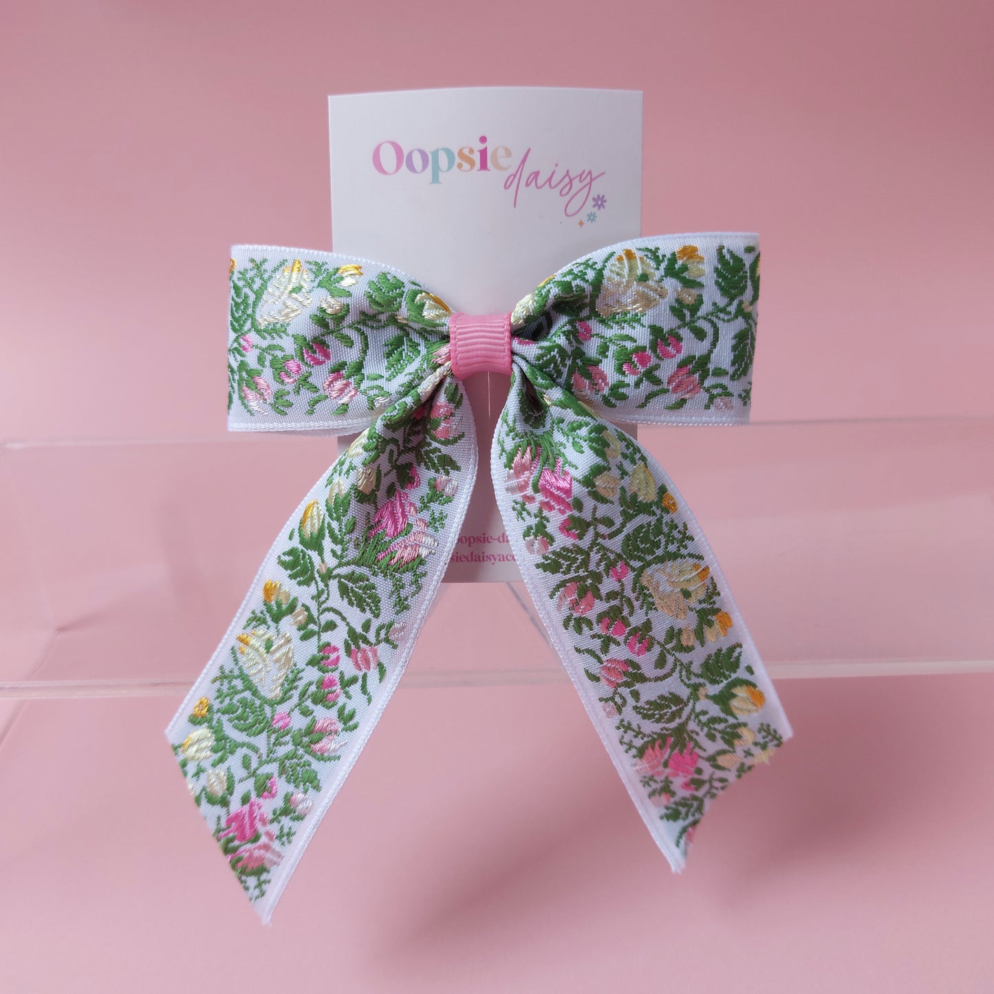 A floral hair bow with green leaves and pink flowers on a white ribbon base, tied with a pink center band, displayed on a pink card that reads 'Oopsie Daisy'.