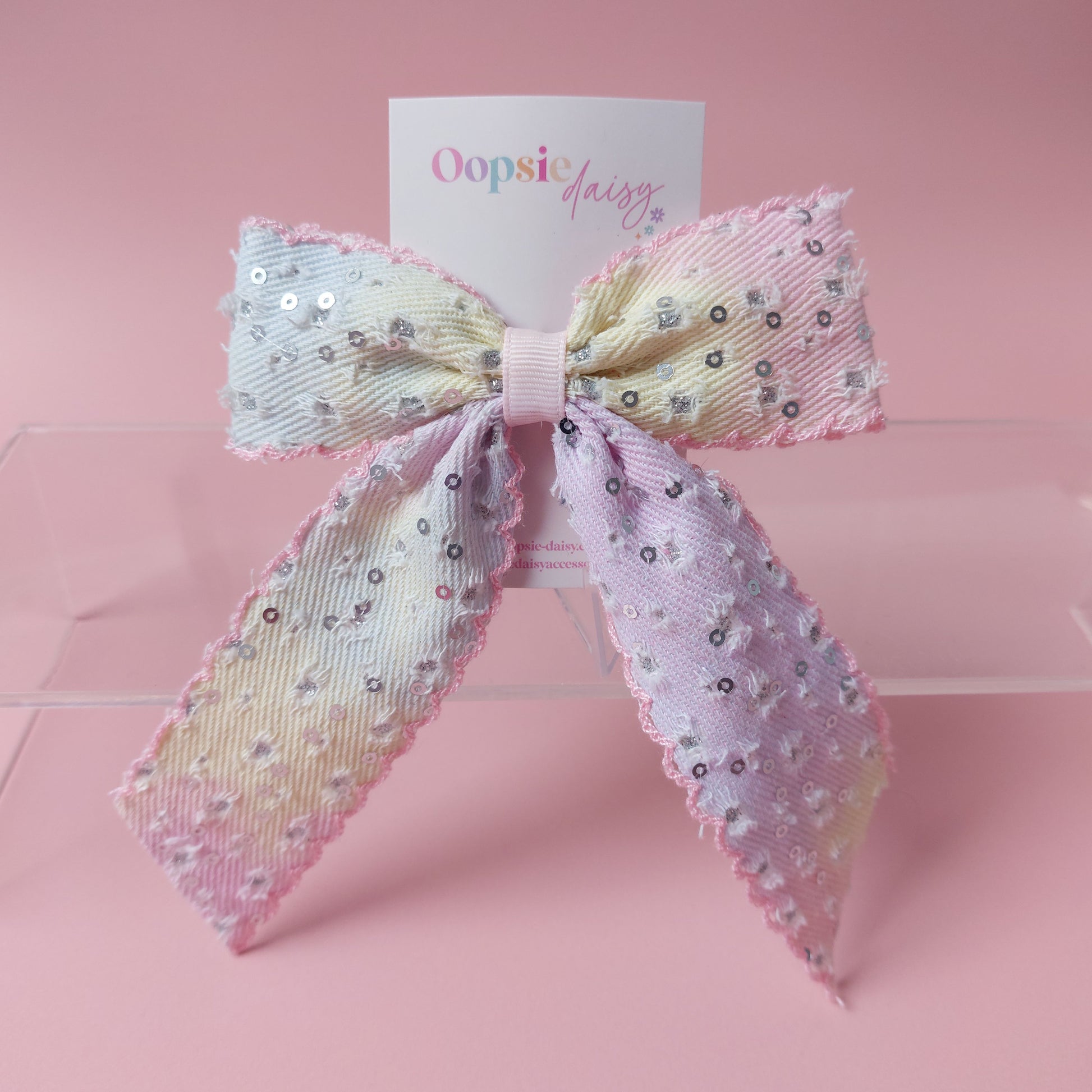 A pastel hair bow bow with sequins, displayed on a clear acrylic stand against a pink background. A white card behind the bow shows the brand name 'Oopsie Daisy'.