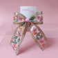 A decorative hair bow with pink floral embroidery, sequins, and metallic gold trim, displayed on a clear acrylic stand against a soft pink background. A card behind the bow reads 'Oopsie Daisy'.
