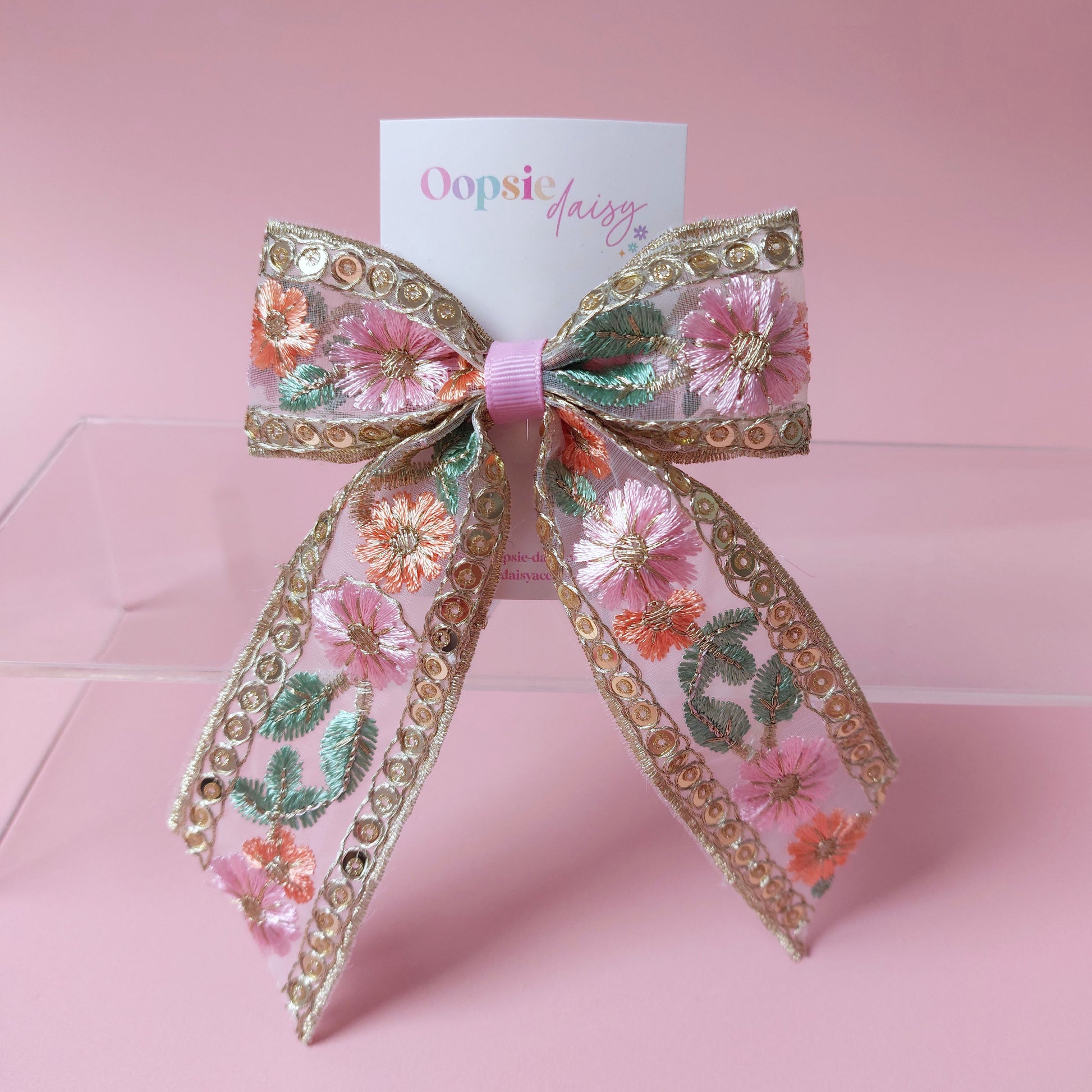 A decorative hair bow with pink floral embroidery, sequins, and metallic gold trim, displayed on a clear acrylic stand against a soft pink background. A card behind the bow reads 'Oopsie Daisy'.