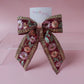 A decorative hair bow with a deep red base, featuring embroidered pink and peach flowers, green leaves, and gold metallic trim. A small white card with the brand name 'Oopsie daily' is tucked behind the bow, which is displayed on a clear acrylic stand against a solid pink background.