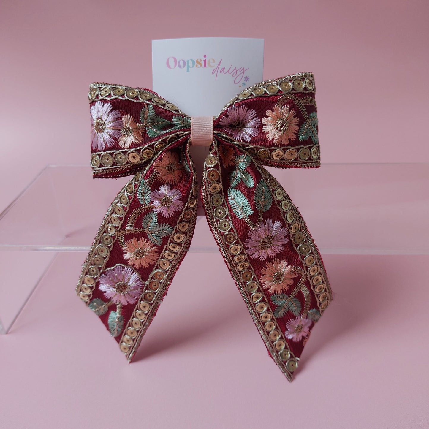 A decorative hair bow with a deep red base, featuring embroidered pink and peach flowers, green leaves, and gold metallic trim. A small white card with the brand name 'Oopsie daily' is tucked behind the bow, which is displayed on a clear acrylic stand against a solid pink background.