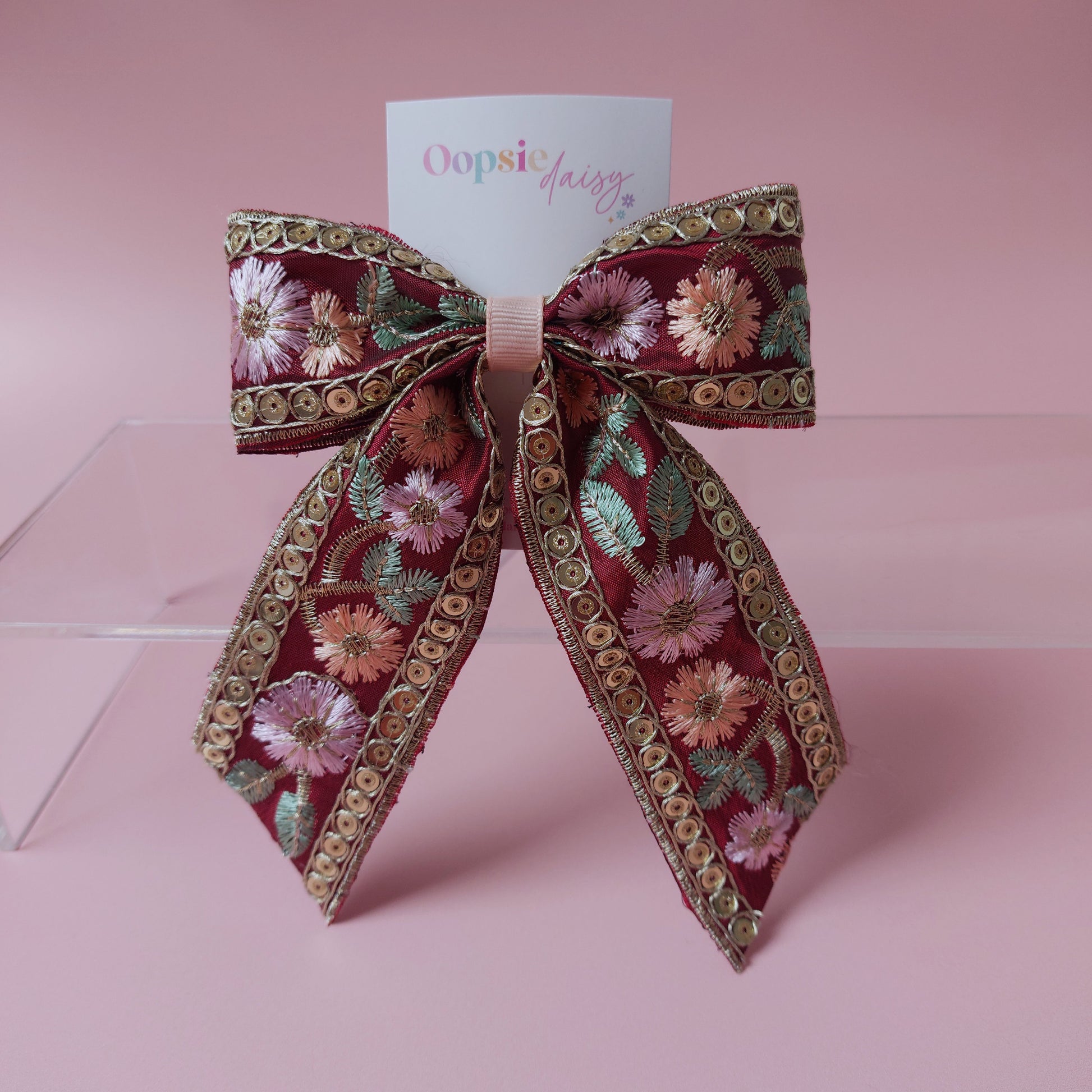 A decorative hair bow with a deep red base, featuring embroidered pink and peach flowers, green leaves, and gold metallic trim. A small white card with the brand name 'Oopsie daily' is tucked behind the bow, which is displayed on a clear acrylic stand against a solid pink background.
