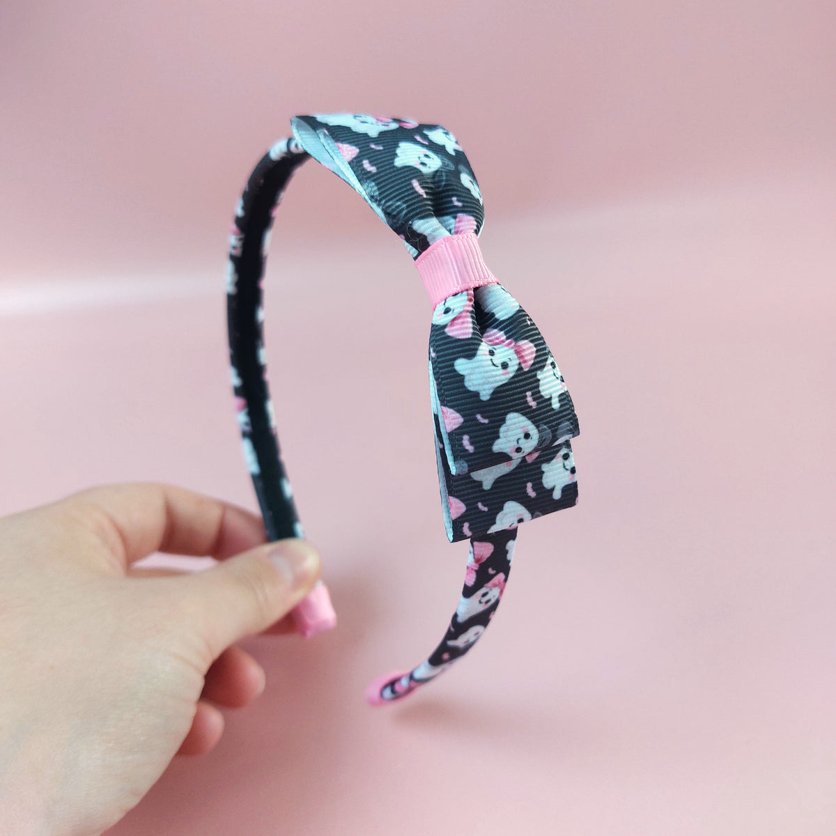 A black alice headband with bow with a pattern of white ghosts wearing pink bows. 