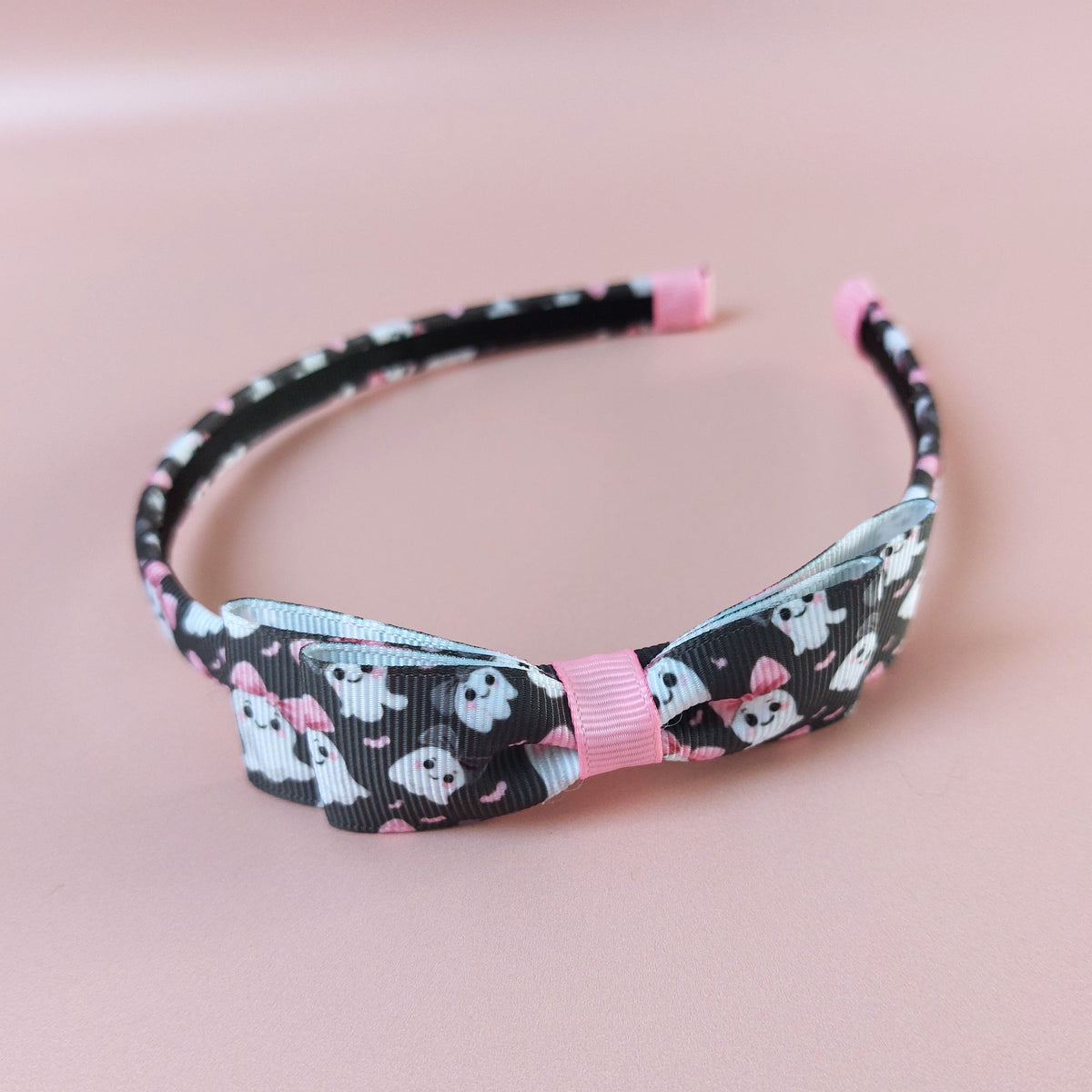 A black alice headband with bow with a pattern of white ghosts wearing pink bows. 