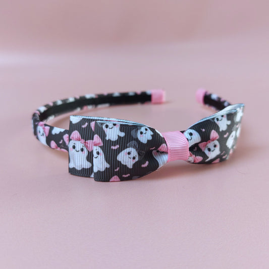 A black alice headband with bow with a pattern of white ghosts wearing pink bows. 