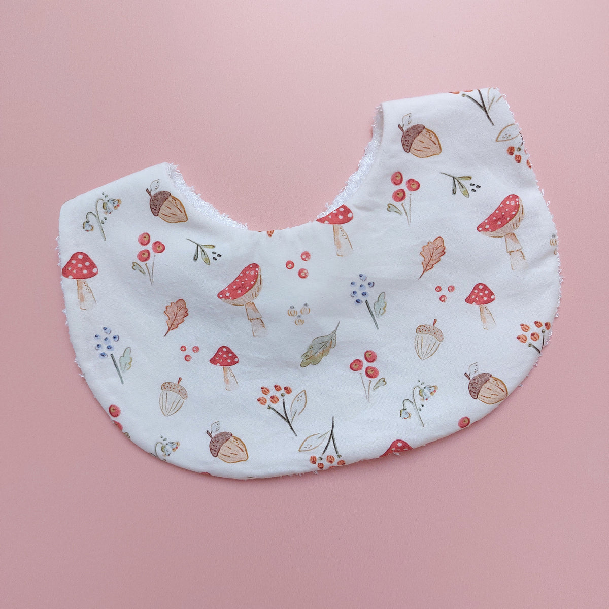 White round shaped baby bib with autumnal pattern of mushrooms, acorns, leaves and autumn flowers. 